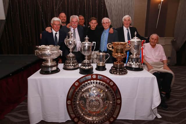 Former Northern Ireland manager Bryan Hamilton (far left) with other Linfield ‘legends’ from the 1970s era – Colin McCurdy, Isaac Andrews, George Dunlop, Eric Bowyer, and Ivan McAllister,  together with current Blues’ star Jordan Stewart, and the seven trophies Linfield won in their ‘clean sweep’ season of 1961-62