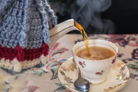 Tea being poured from a vintage bone china tea pot with with knitted wool cosy or cozy cover into a pretty tea cup with saucer. All on a vintage style patterned table cloth. Steam can be seen coming from the hot drink.