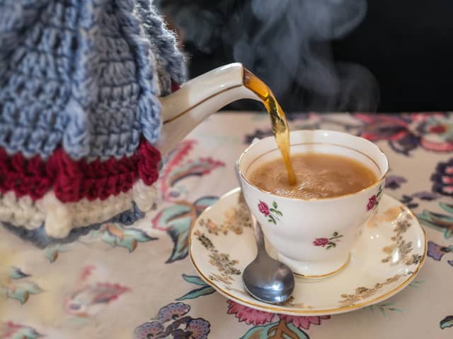 Tea being poured from a vintage bone china tea pot with with knitted wool cosy or cozy cover into a pretty tea cup with saucer. All on a vintage style patterned table cloth. Steam can be seen coming from the hot drink.