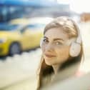 Young woman with freckles listens to music through white headphones, enjoying a sunny city vibe.