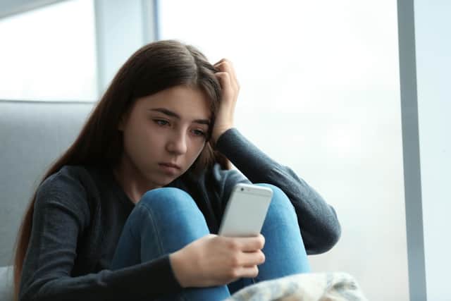 Upset teenage girl with smartphone sitting at window
