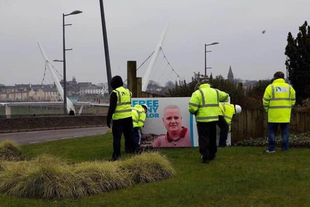 A photograph showing a group of men, some of whom are wearing Derry City and Strabane District council branded clothing, erecting the 'free Tony Taylor' poster at the roundabout at Harbour Square in Londonderry city centre